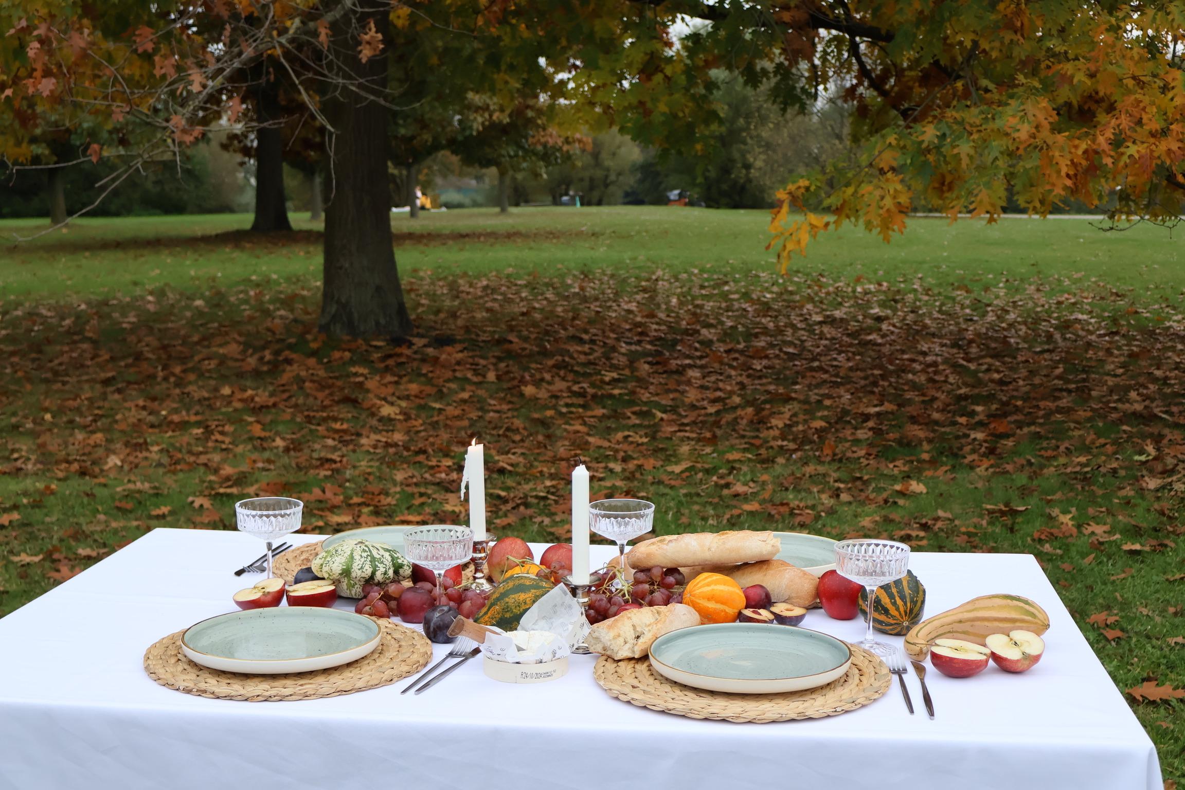 Ein kreativ dekorierter Tisch mit Obst, Gemüse, Kerzen, Sektgläsern und Käse steht draußen im Park an einem Herbsttag.